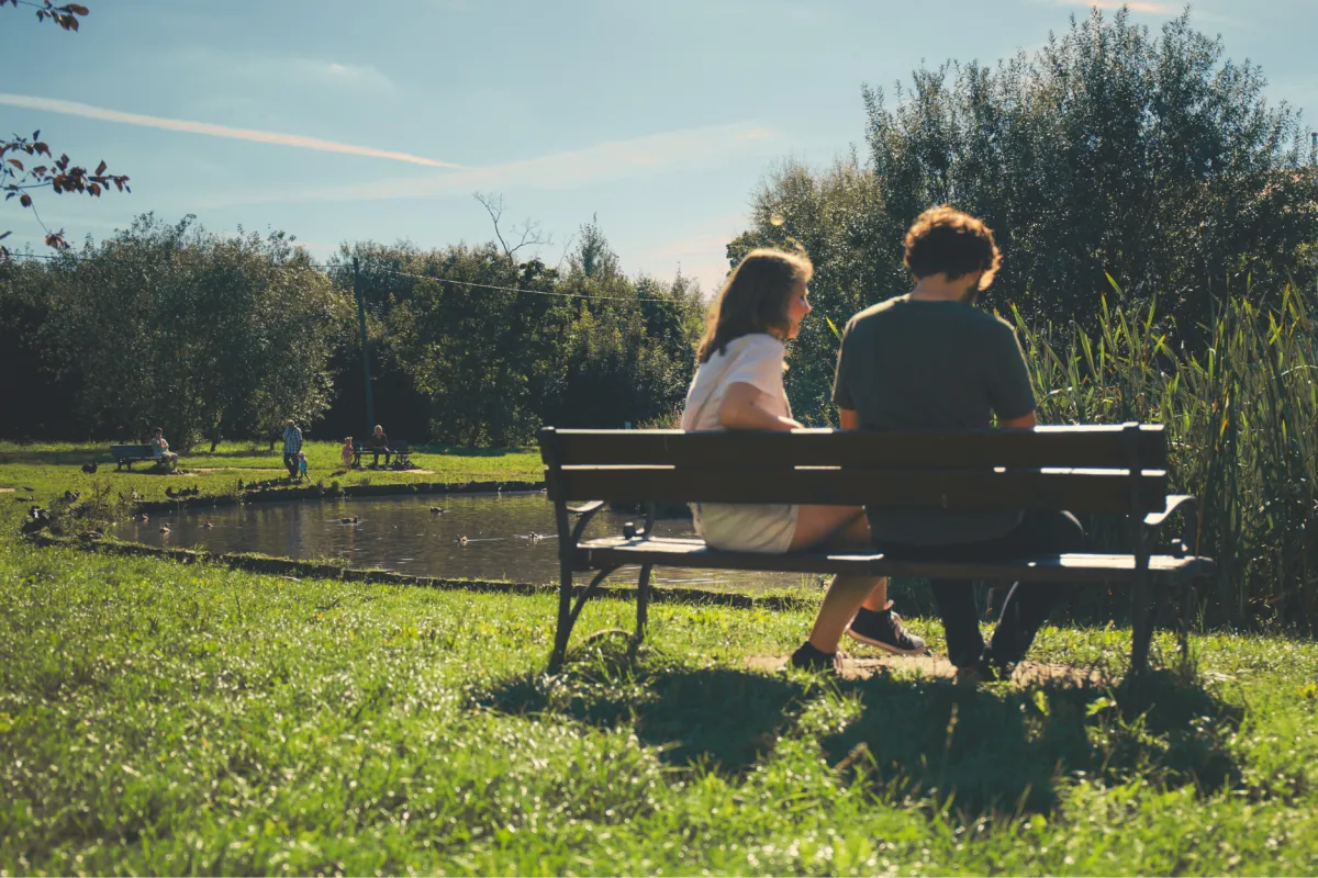 Two people sitting on a bench by a pond in a park, talking in a calm outdoor setting.