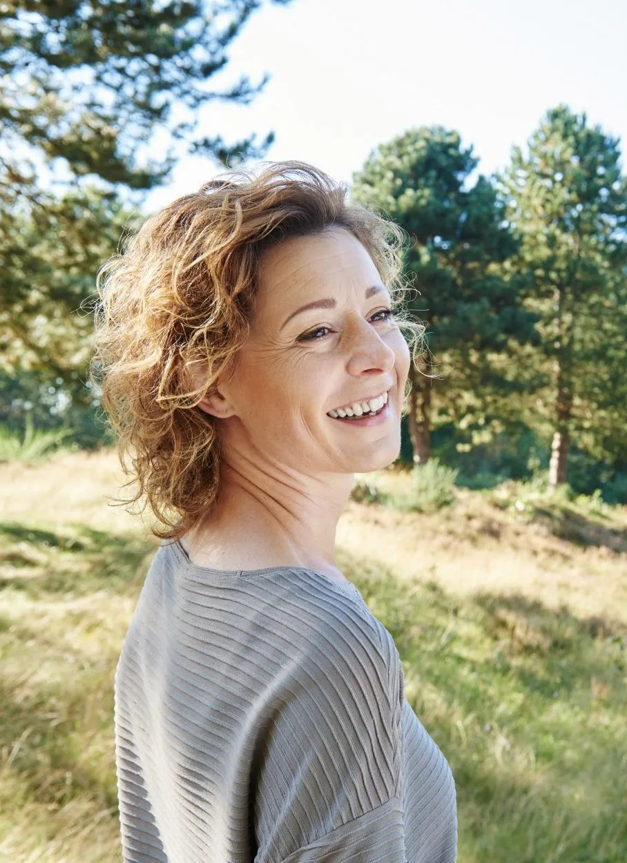 Woman with curly hair smiling in a sunlit forest, representing joy and connection in nature