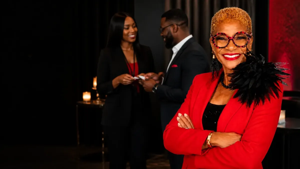 Black woman networking and sales coach in a red blazer smiling confidently while two business professionals exchange contact information at a networking event.