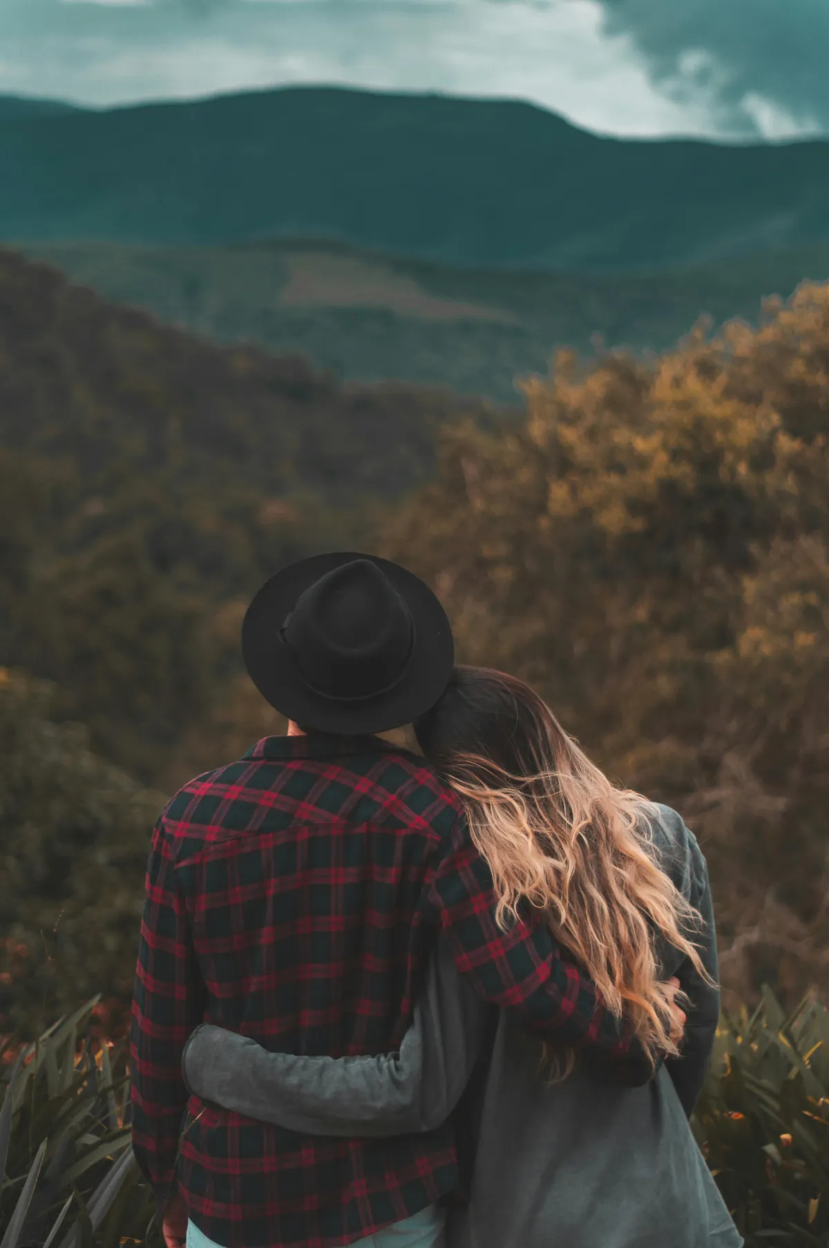 Two people with their backs facing the camera, embracing as they look at scenic view of mountains and greenery