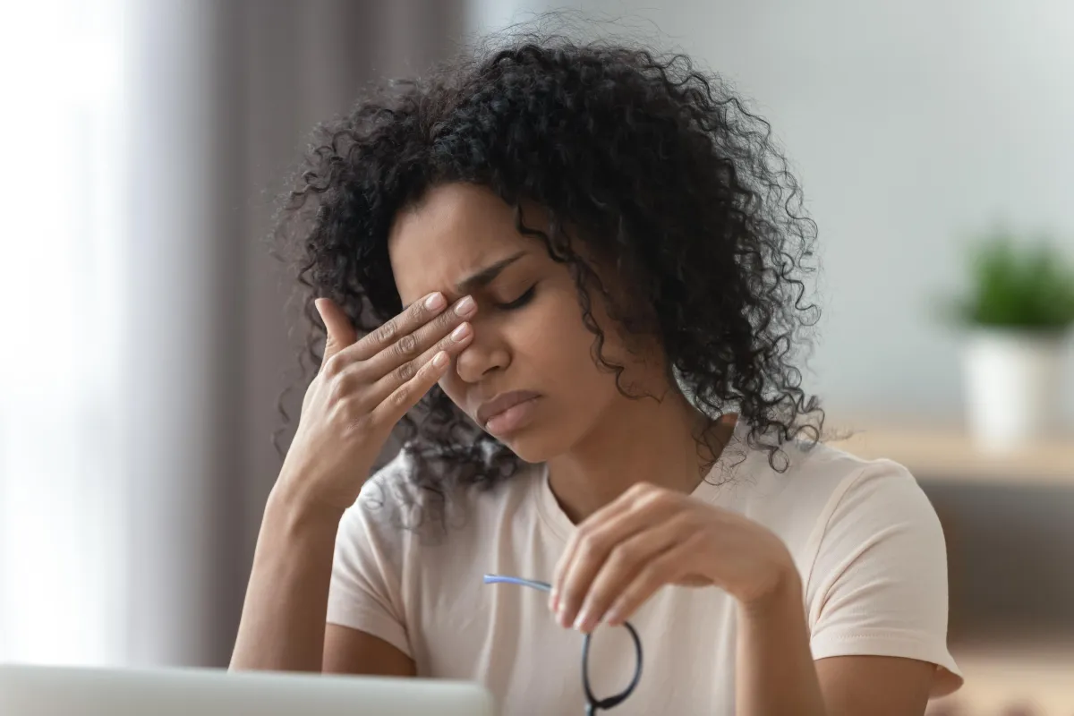 Black woman with curly hair holding her head in frustration while working on a laptop, symbolizing fatigue and chronic illness burnout.
