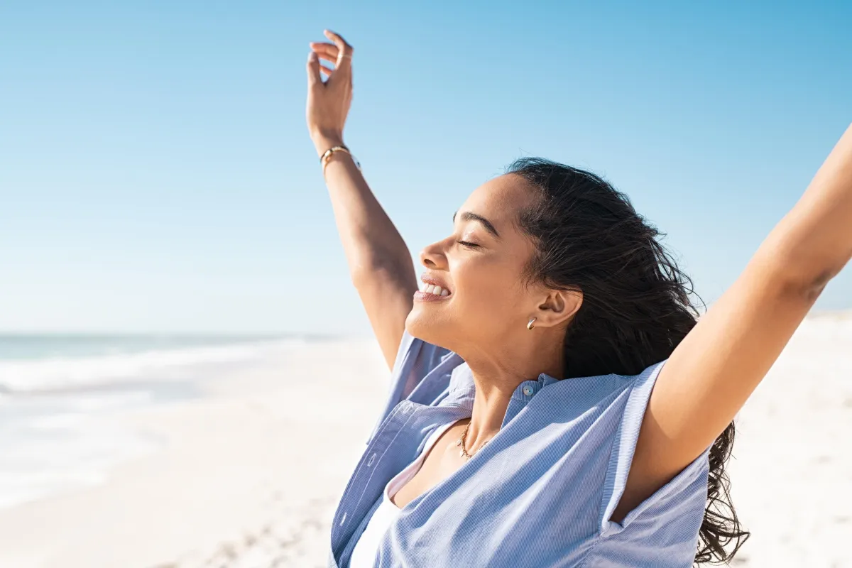 Joyful hispanic woman stretching arms toward the sky on a beach, symbolizing energy, healing, and empowerment.
