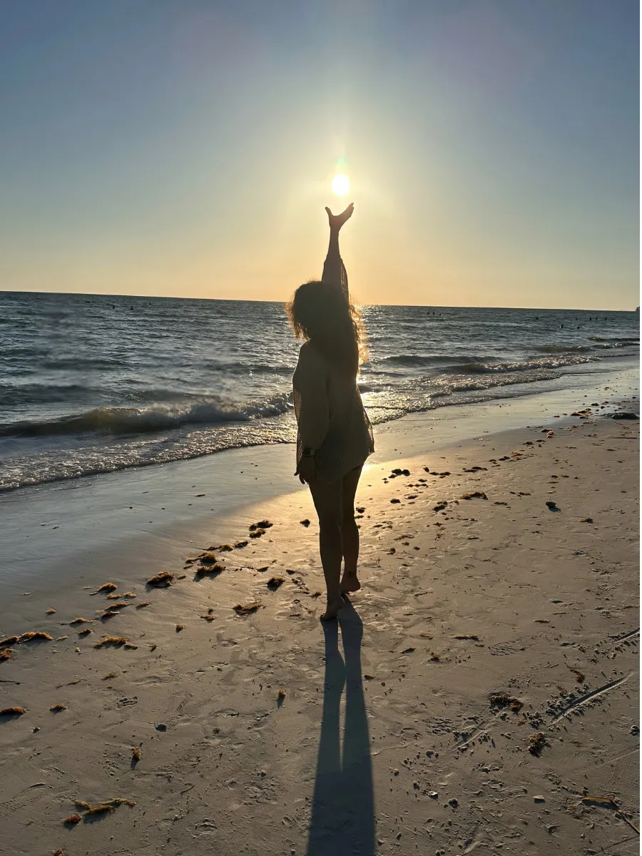 Silhouette of a woman standing on the beach at sunset, reaching toward the sun with one hand—symbolizing peace, clarity, and empowered transformation.