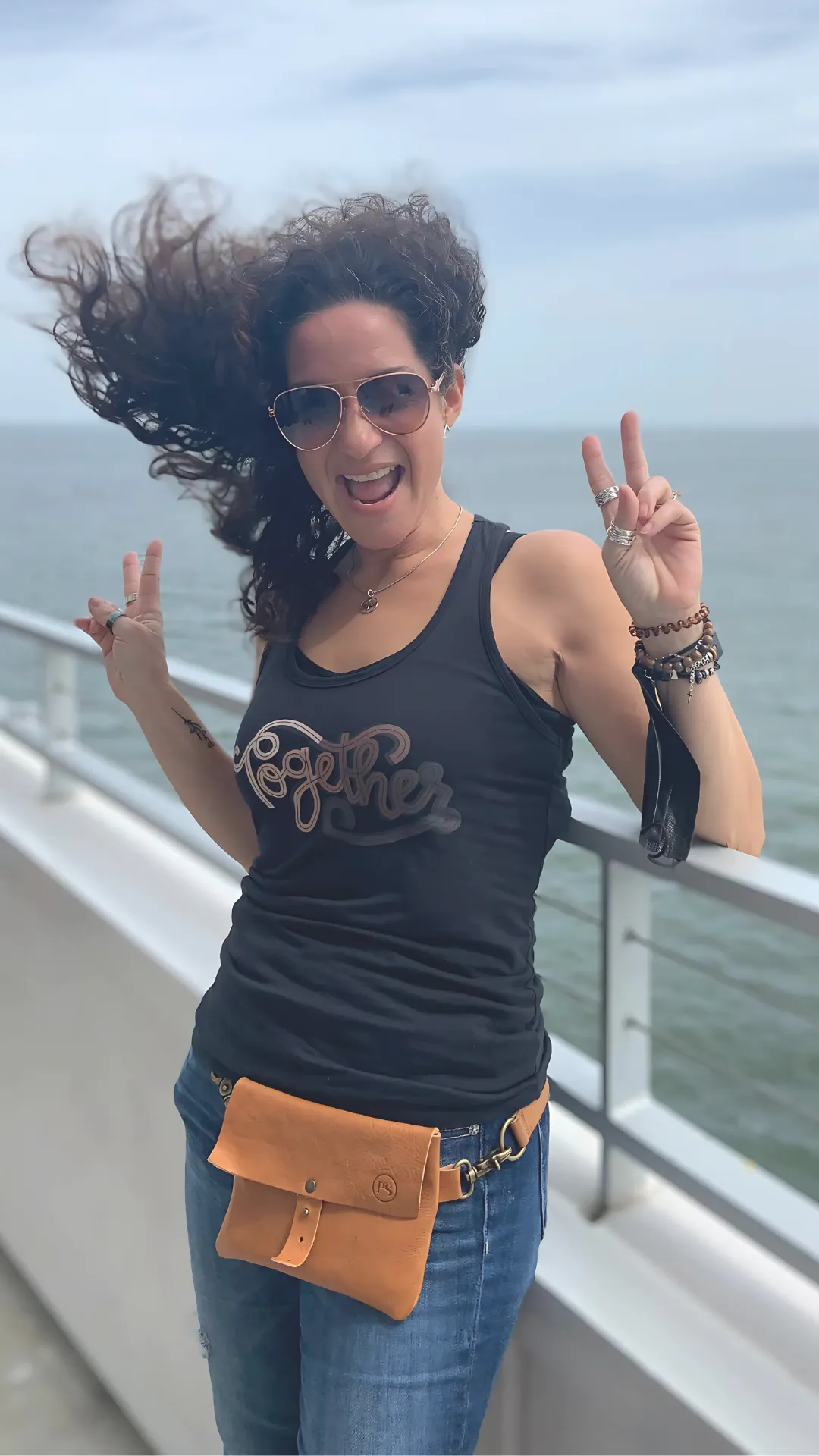 Smiling woman on a breezy oceanfront deck, throwing peace signs with windblown hair—capturing joy, energy, and carefree self-expression.