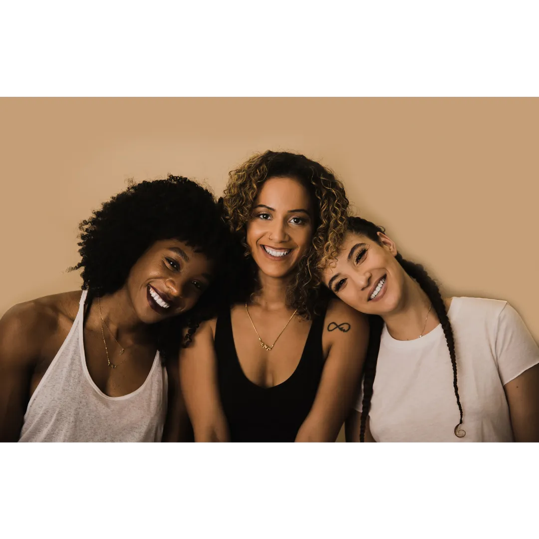 Three diverse women smiling and leaning on each other against a neutral background, symbolizing community, connection, and empowered sisterhood.