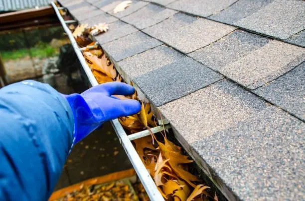 Home maintenance service by Home Star Roofing, with a gloved hand removing leaves and debris from a clogged gutter to prevent water damage.