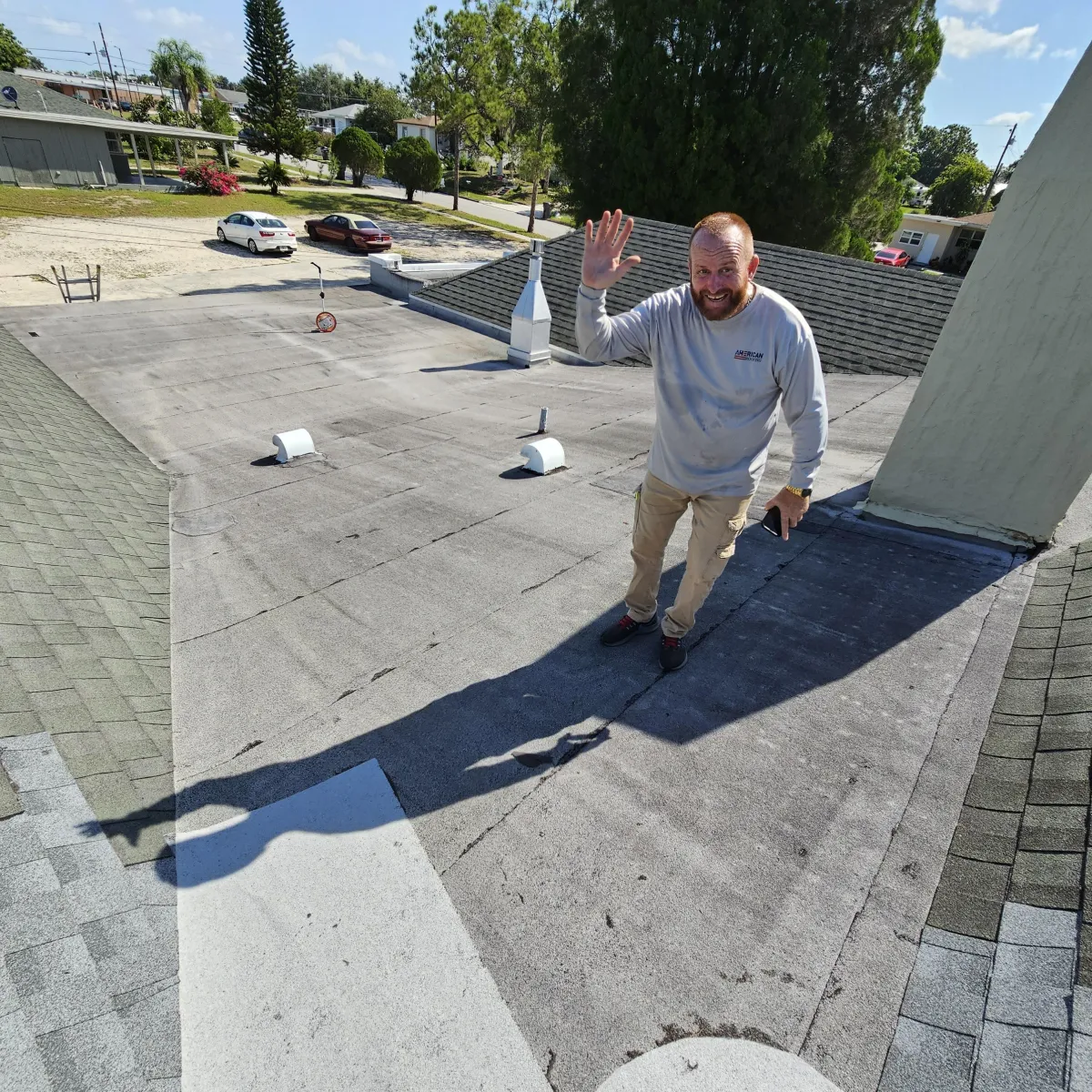 American Roofing FL technician waving during flat-roof inspection; vents and parapet visible.