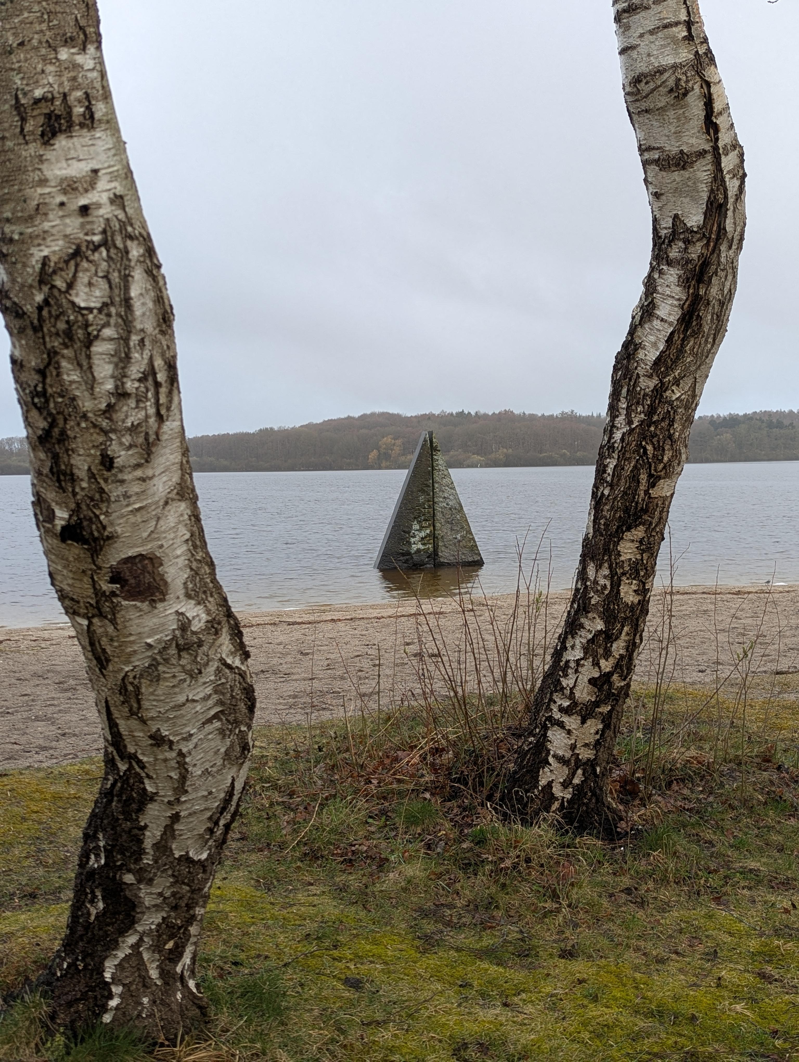 A scenic picture looking between two tree trunks over a lakeside beach and still water with a stone pyramid sculpture in the water.