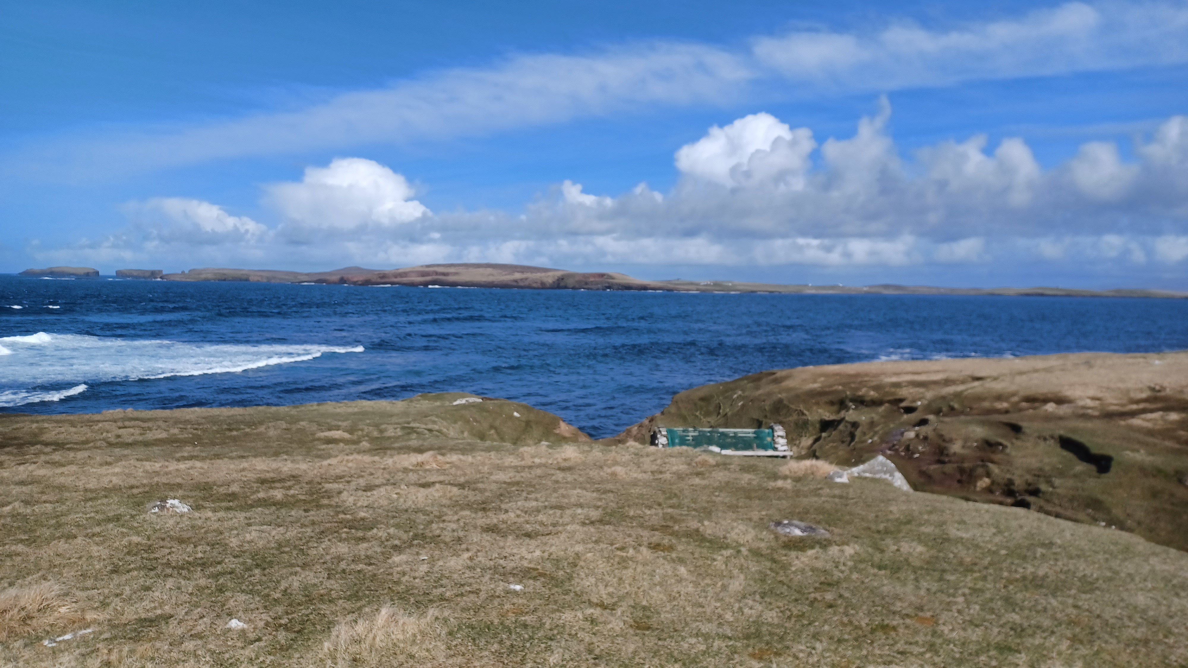 A view from Shetland on a bright sunny day with an active sea, an island in the distance and white clouds in the blue sky.