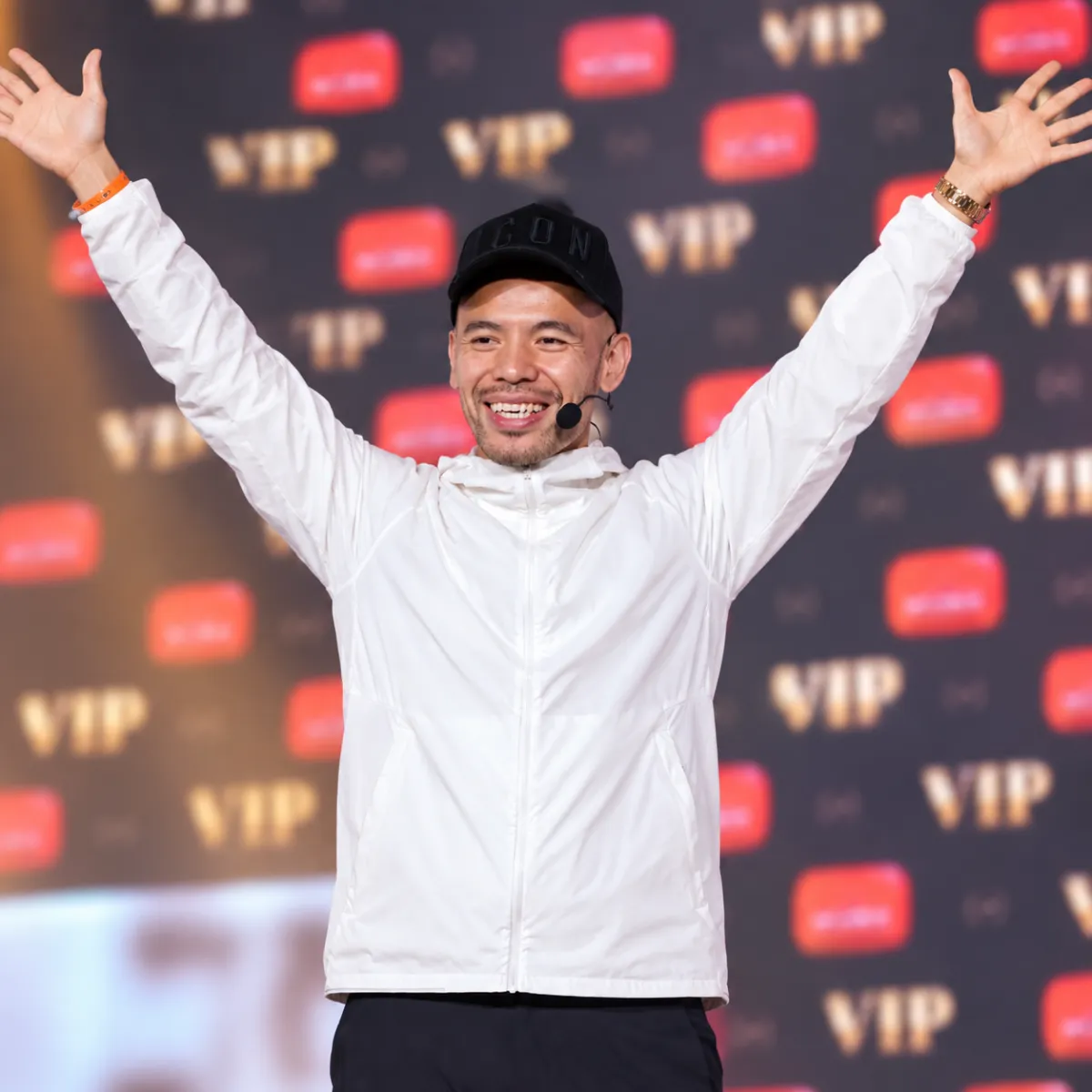 Smiling Asian man holding a golden YouTube play button plaque, wearing a blue shirt and black cap