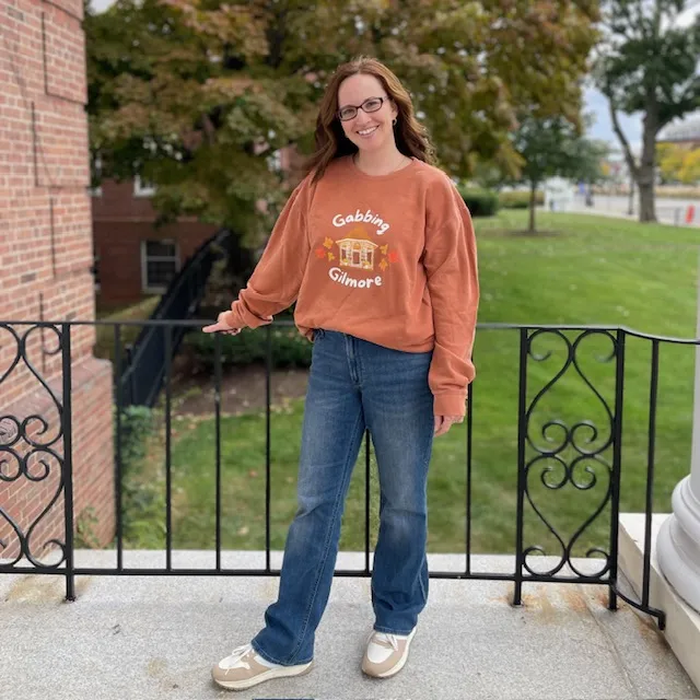 catherine standing with an orange gabbing gilmore sweatshirt and blue jeans with trees and grass in the background