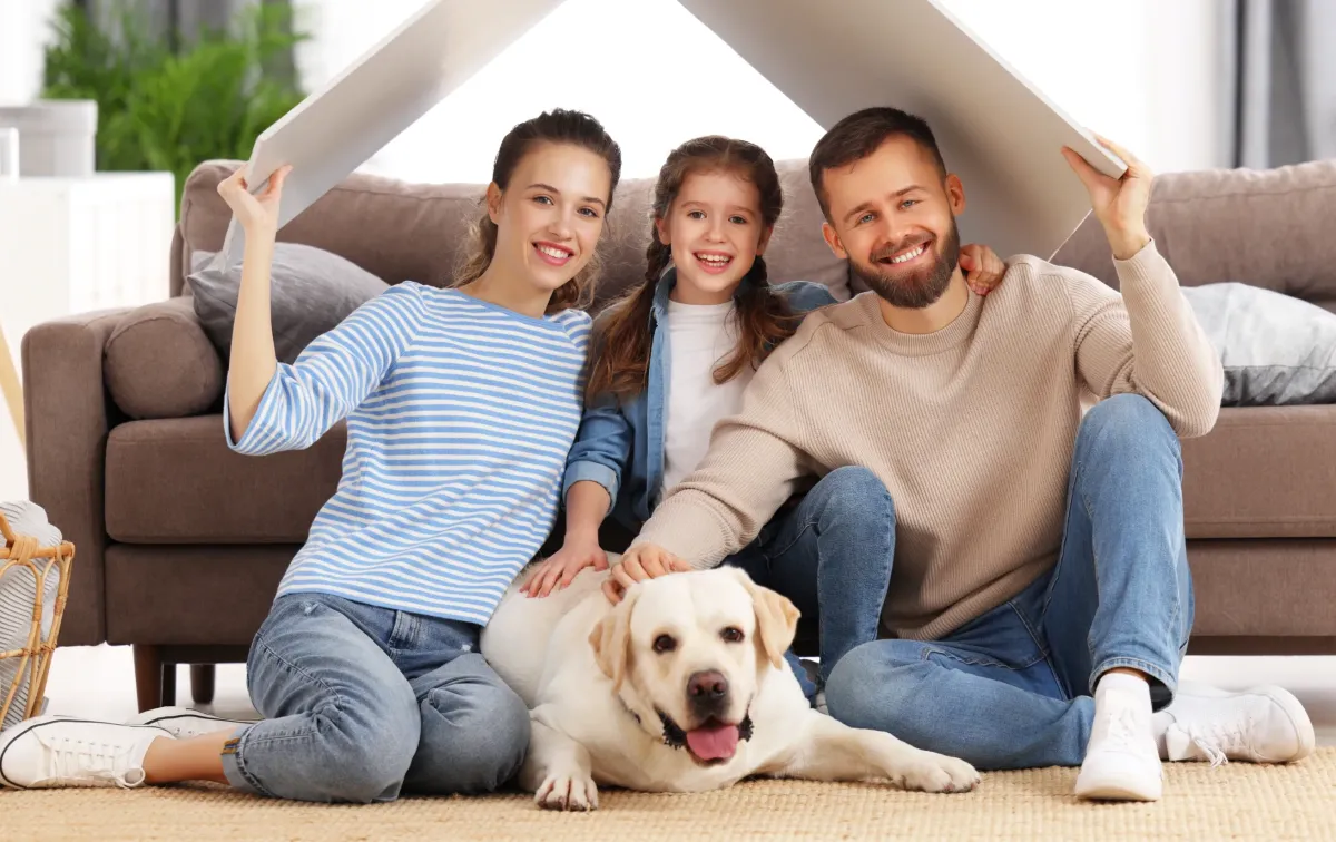 Smiling family with young daughter and Labrador dog sitting in cozy living room, symbolizing comfort and safety in a new home.