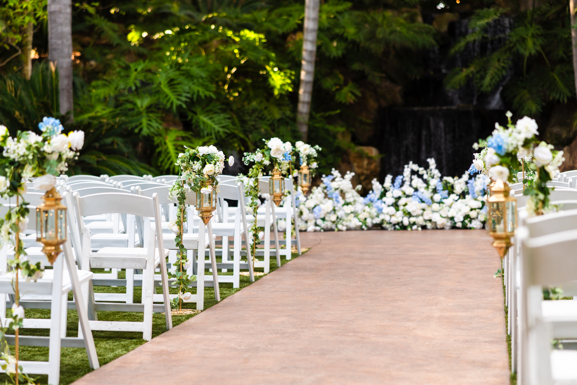 Outdoor wedding ceremony aisle lined with white folding chairs, tall gold lanterns wrapped in eucalyptus garlands, and white and blue hydrangea floral arrangements leading toward a waterfall backdrop at Grand Tradition Estate in Fallbrook.