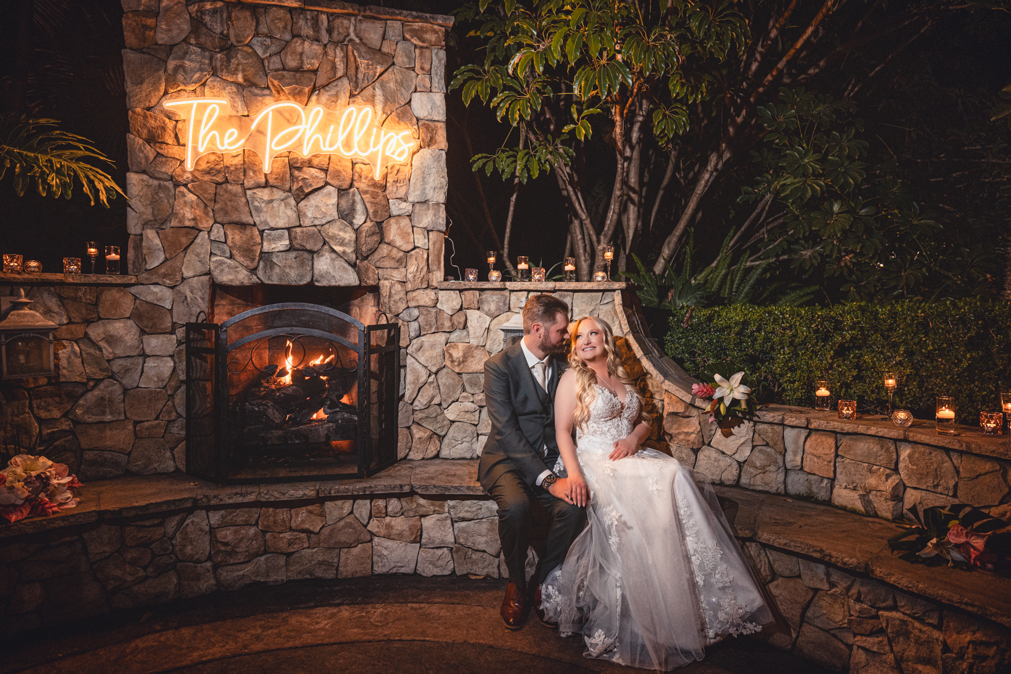 Bride and groom seated together on stone steps in front of a lit outdoor fireplace at night, with a glowing neon sign reading "The Phillips" mounted on the stone chimney, surrounded by candles and tropical greenery at Grand Tradition Estate.