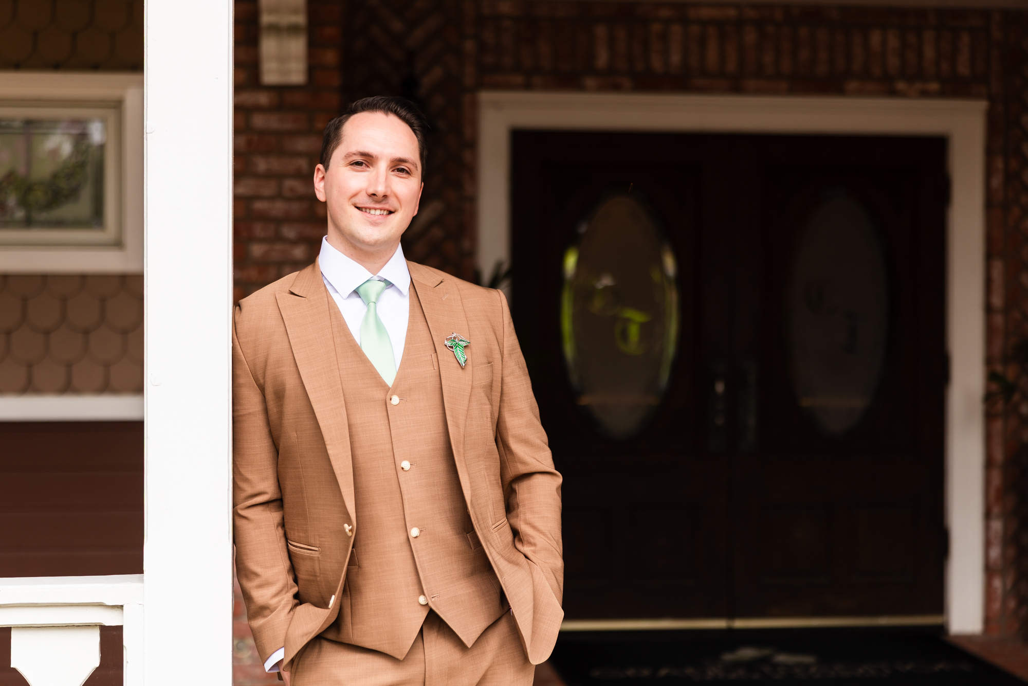 Groom in a tan three-piece suit with a mint green tie and decorative lapel pin, smiling while leaning against a white column in front of a brick building with a dark arched door.