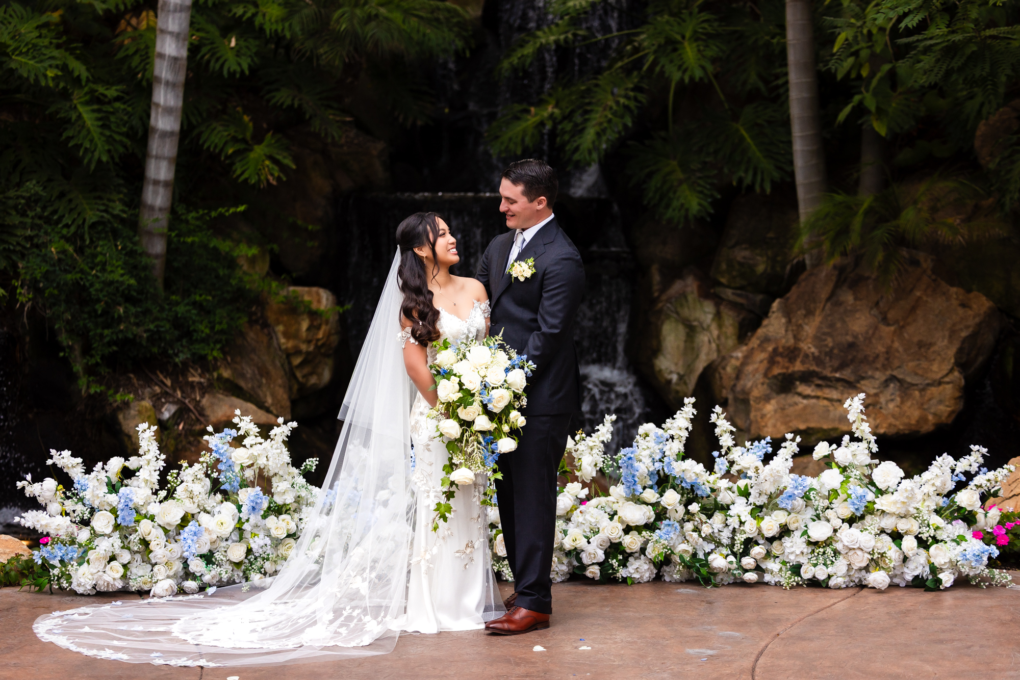 Bride and groom standing together in front of a cascading waterfall altar framed by an abundant low floral arrangement of white roses and blue hydrangeas at Grand Tradition Estate in Fallbrook, California.