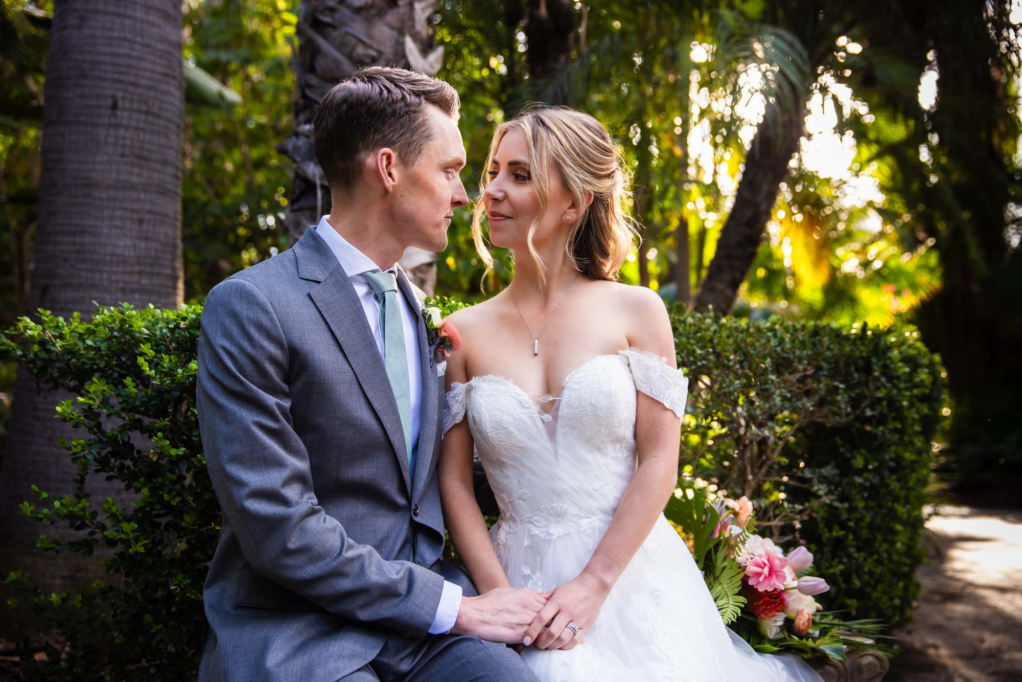 Bride and groom seated close together on a garden bench, looking at each other in soft golden hour light filtered through palm trees and lush hedges at Grand Tradition Estate, the bride holding a tropical floral bouquet.