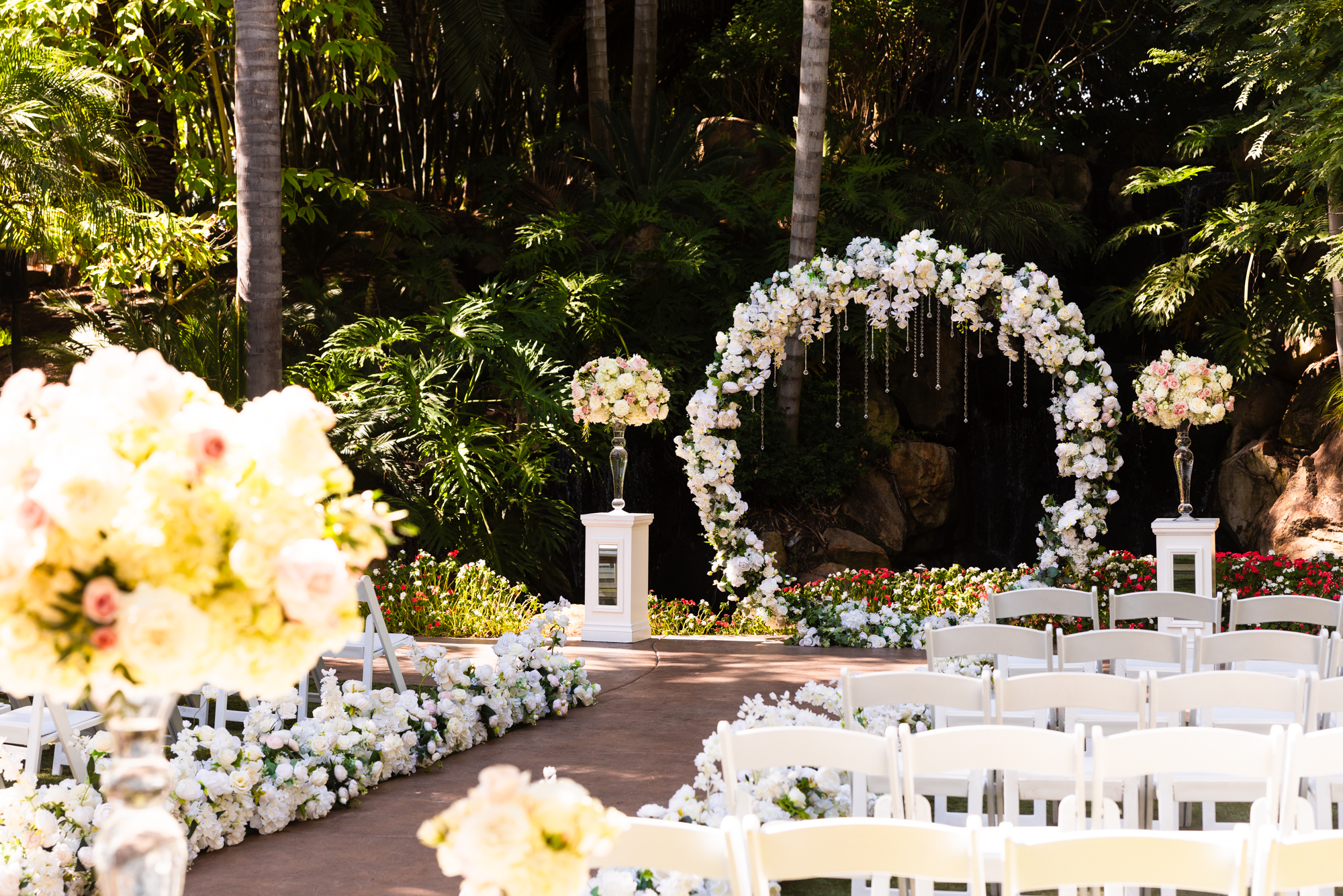 Outdoor wedding ceremony setup at Grand Tradition Estate featuring a full white floral arch with crystal accents, flanked by tall floral pedestals, with white chairs lining a petal-covered aisle and tropical greenery in the background.