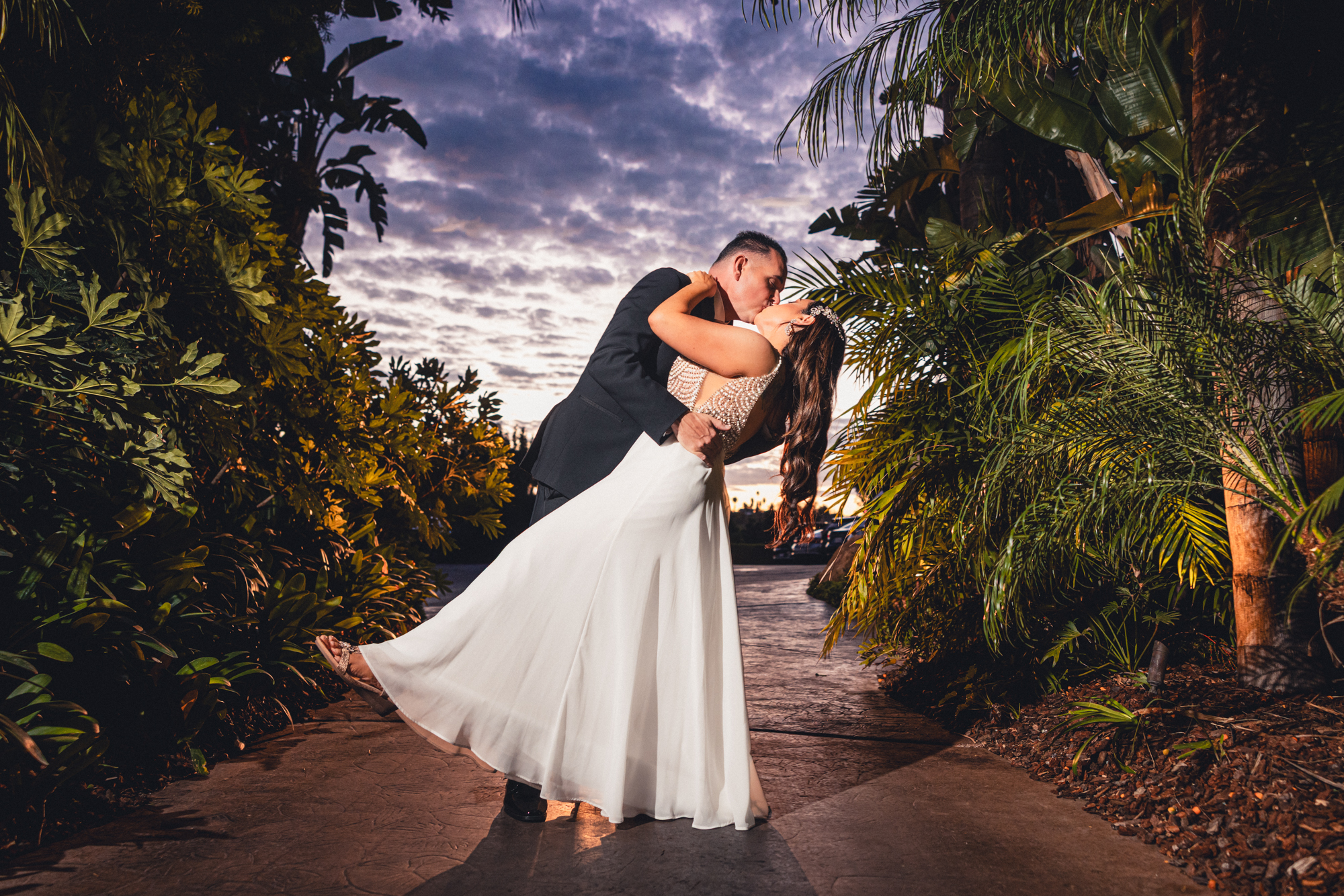 Bride and groom sharing a dramatic dip kiss at twilight along a palm-lined garden path at Grand Tradition Estate, with a vivid purple and orange cloudy sky visible above the tropical foliage.