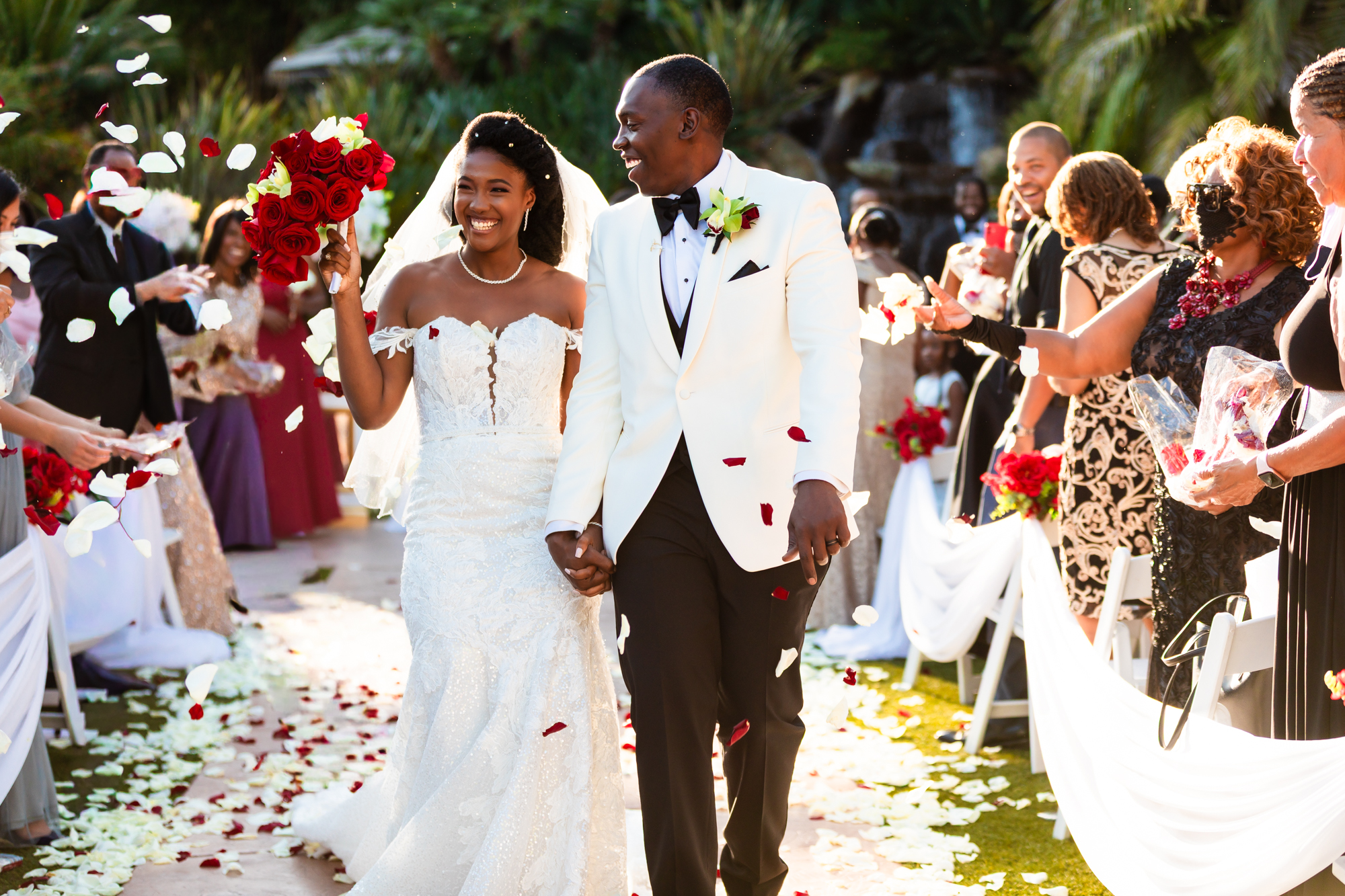 Bride and groom walking back up the aisle after their ceremony at Grand Tradition Estate, laughing joyfully as guests toss red and white rose petals around them, the bride in a fitted off-shoulder lace gown holding a bouquet of red roses and the groom in a white tuxedo jacket with black bow tie.