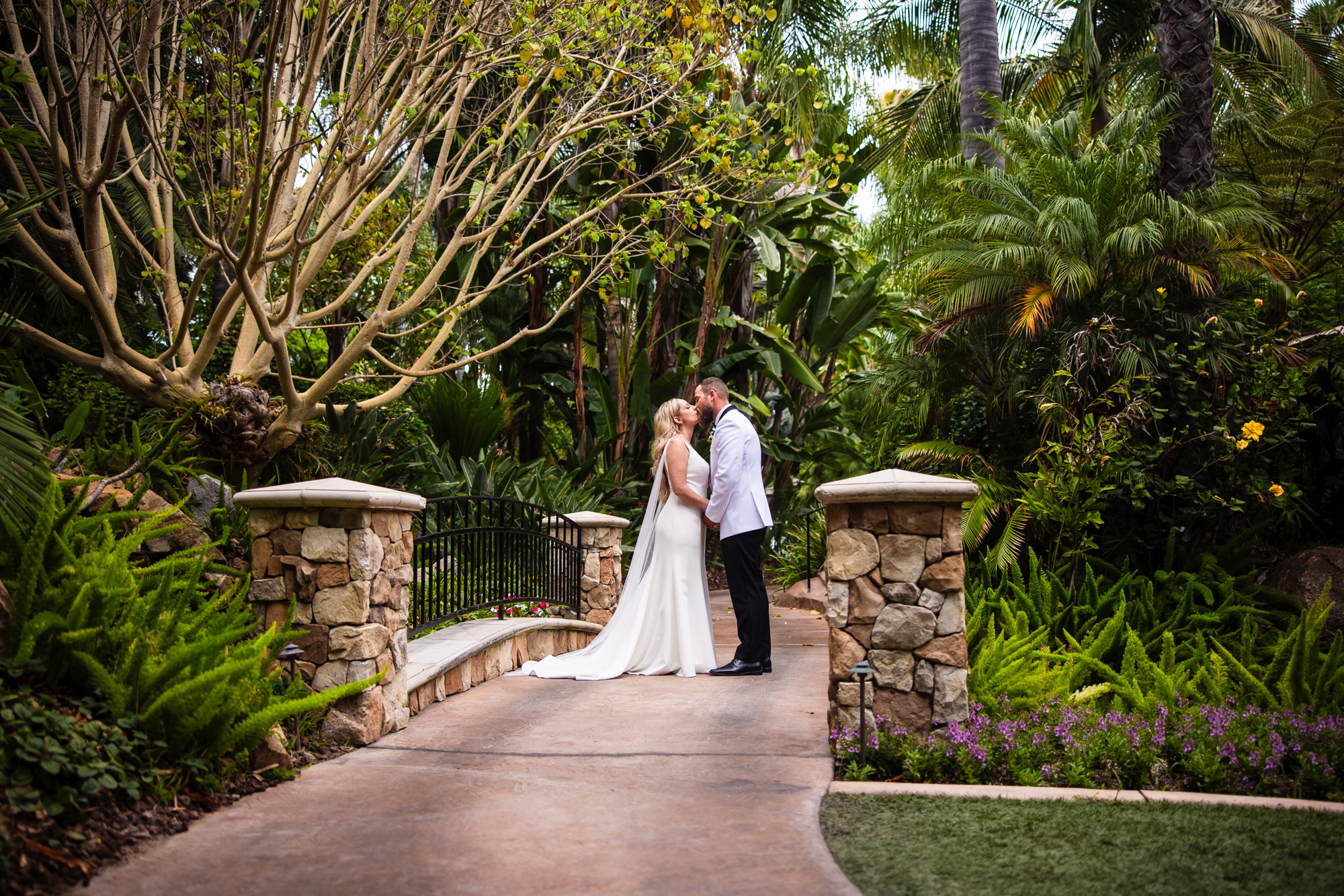 Bride and groom sharing a kiss on a stone-pillared garden bridge at Grand Tradition Estate, surrounded by dense tropical foliage, palm trees, ferns, and flowering purple ground cover, the bride's cathedral veil trailing behind her on the path.