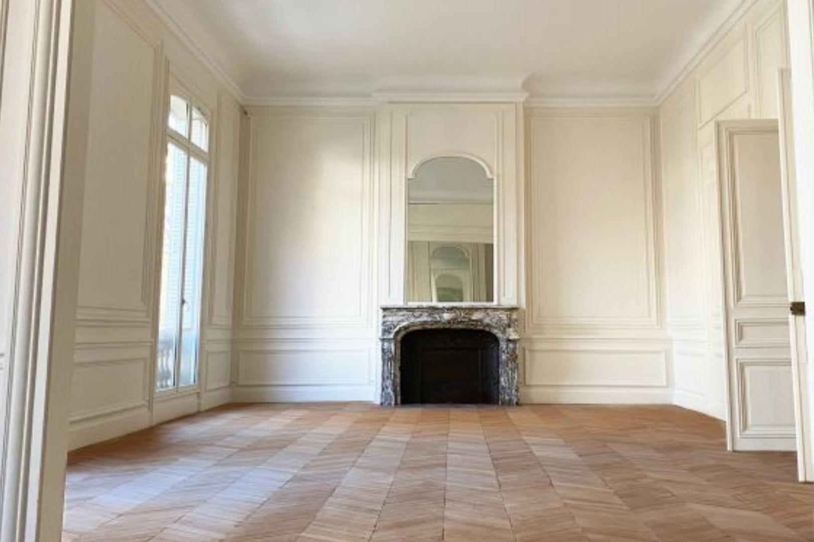 Grand salon rénové avec parquet en chevrons, moulures aux murs et cheminée ancienne restaurée.