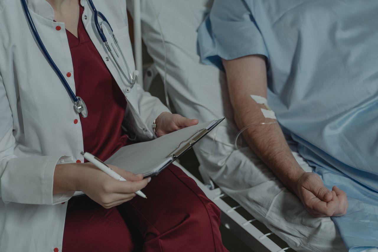 A doctor sits beside a patient in a hospital room, taking notes.