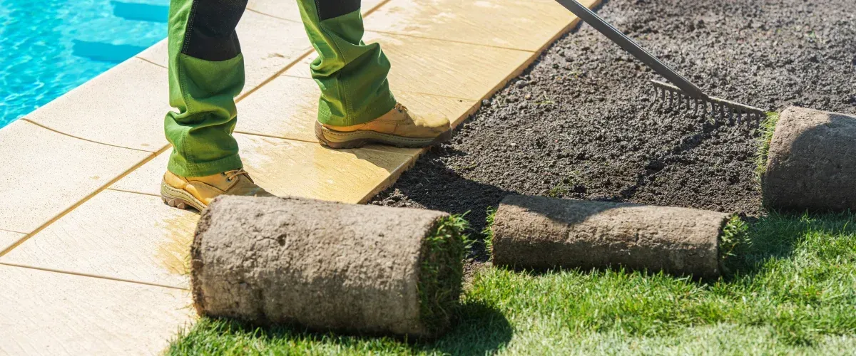 A gardener in green trousers and work boots lays rolls of sod beside a swimming pool on a sunny day.