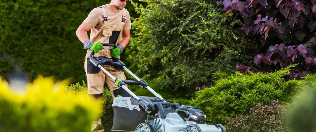 A man in tan overalls and green gloves operates a modern lawnmower in a lush green and purple garden.