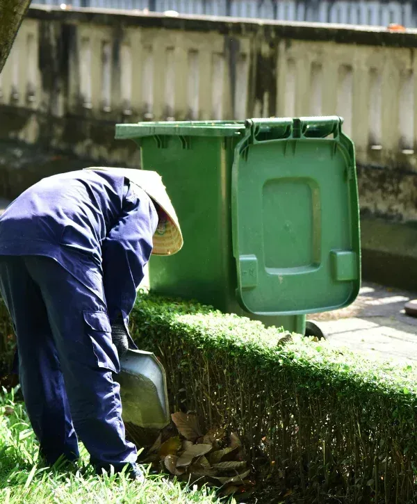 A person in blue work clothes and a conical hat raking leaves into a pile beside a green bin.
