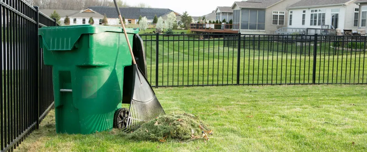 Gardening tools including a rake, hand fork, pruning shears, and gloves laid on brown fallen leaves.