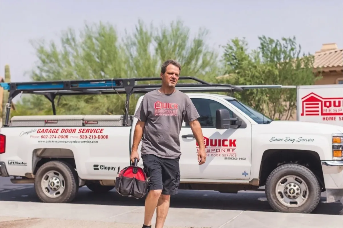 A technician wearing a 'Quick Response Garage Door Service' shirt carries a tool bag, standing in front of his service truck.