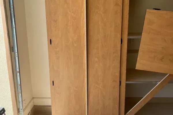 A light brown wooden sliding cabinet door that has fallen off its track, revealing interior shelves.