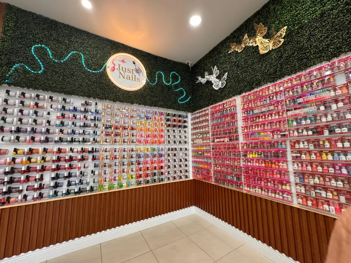 Exterior of Just Nails By Karla salon, featuring a welcoming entrance with large glass doors, pastel signage, and potted plants. The setting is bright and inviting, suggesting easy access for families and professionals.