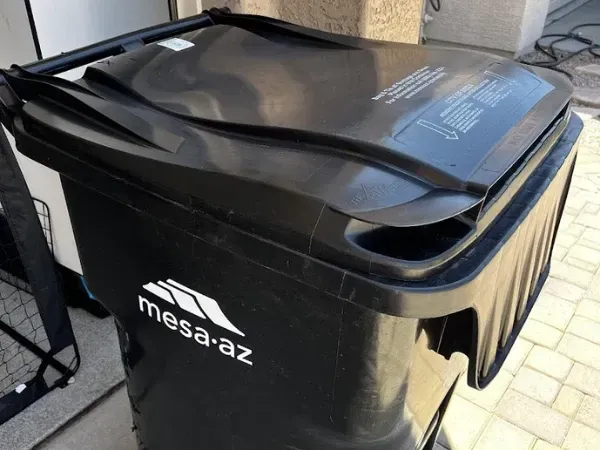 A black residential trash bin provided by Mesa, AZ, showing its closed lid and wheels for curbside waste collection.