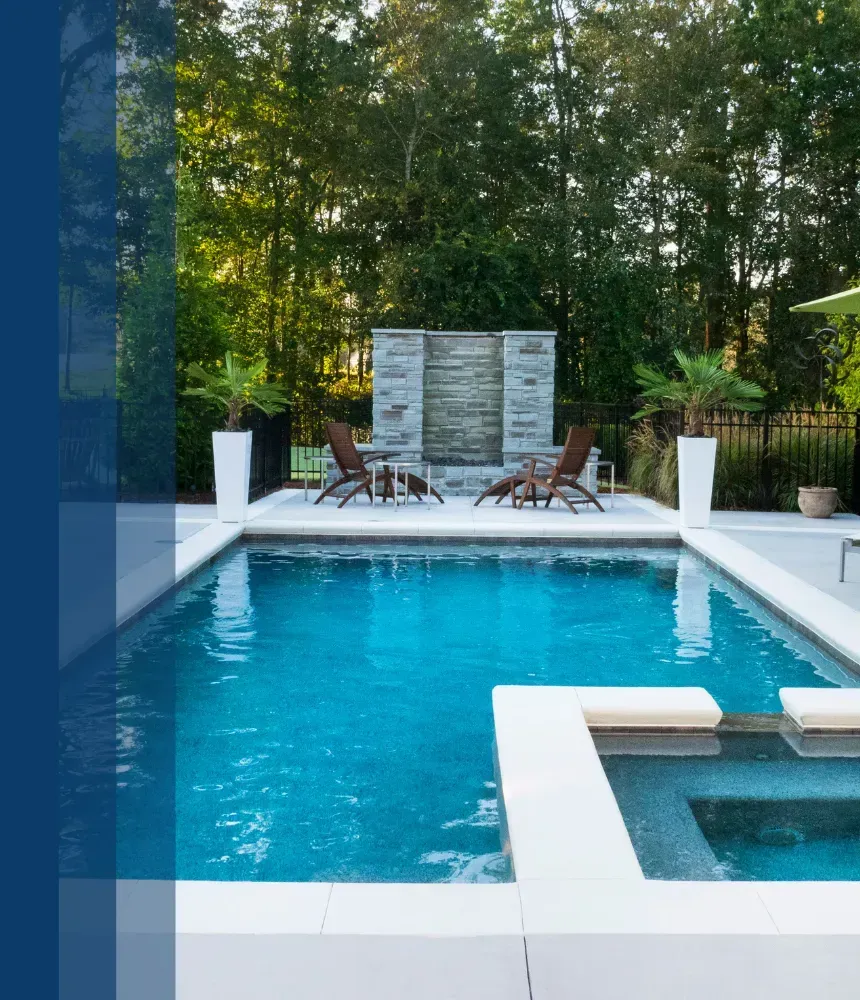 Modern rectangular swimming pool featuring a stone water feature, lounge chairs, and tall trees in the background.