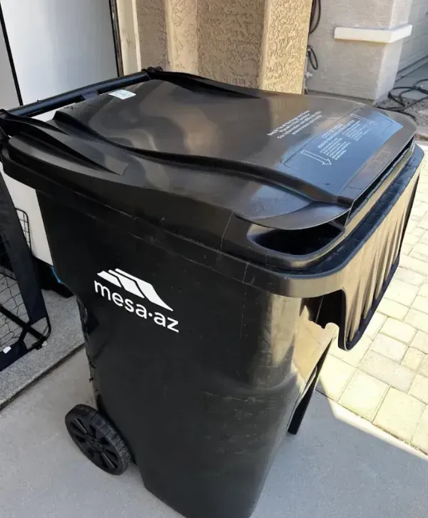 Black residential trash bin with Mesa, Arizona city logo, placed beside a house garage.