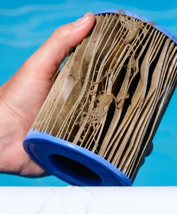 Close-up of a hand holding a dirty and damaged pool filter cartridge with debris caught in the pleats, showing the need for replacement.