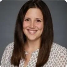 A smiling woman with long brown hair wearing a white and grey patterned blouse