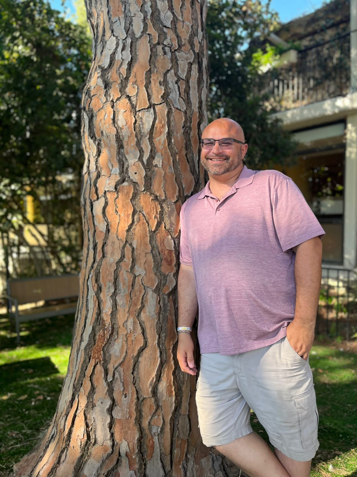 Smiling man in a pink polo shirt and khaki shorts leaning against a large pine tree in a sunny park, with greenery and a residential building in the background.