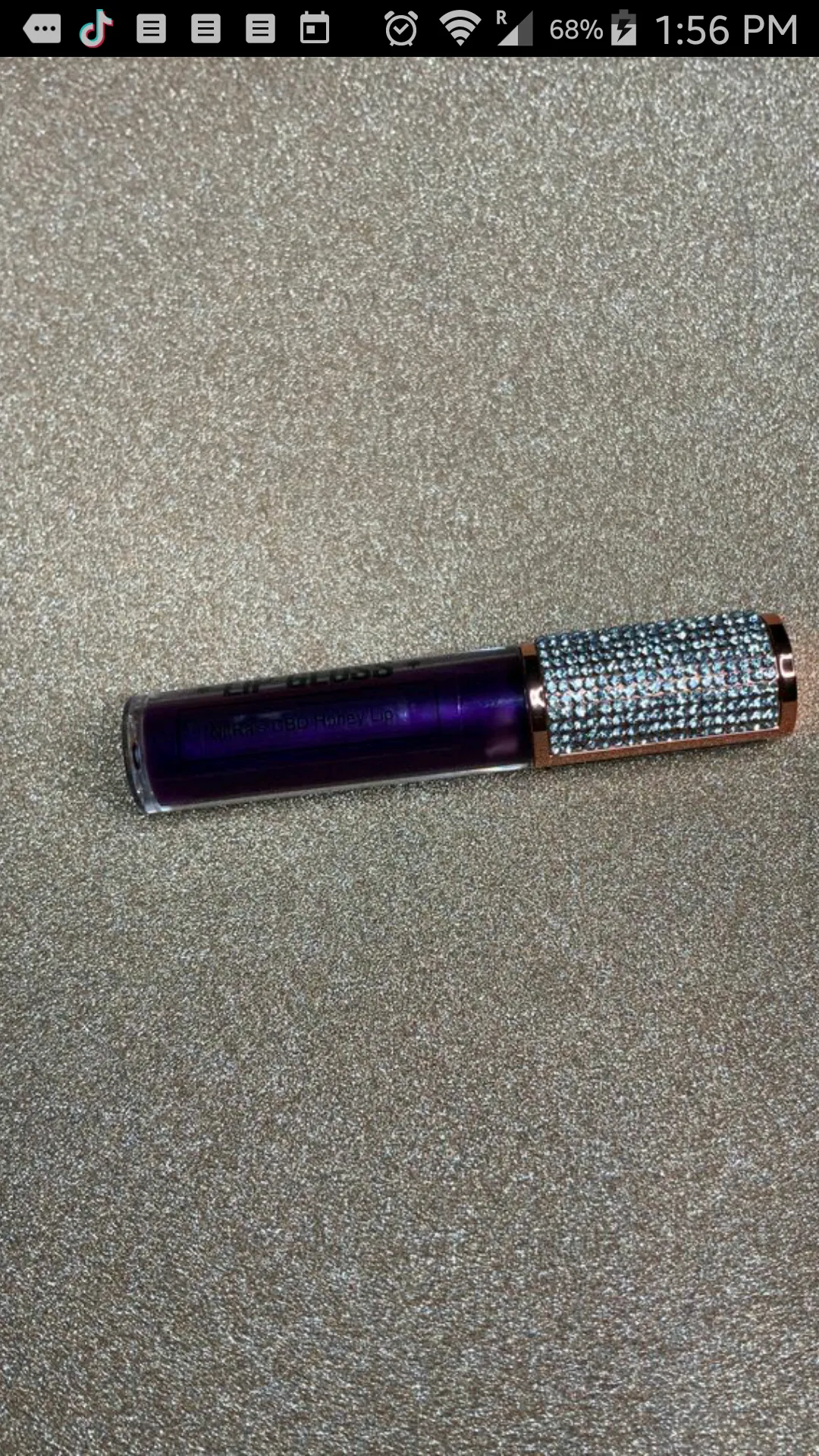 Close-up of a deep purple lip gloss tube with a rhinestone-studded cap, labeled “NitRa’s CBD Honey Lip,” placed on a textured gold background. 