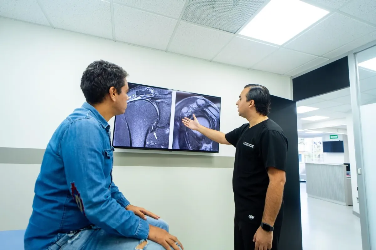 A man standing indoors next to a whiteboard is pointing at an x-ray of another man's body. The setting includes clothing and a ceiling, with a wall in the background.
