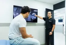 A man wearing casual clothing is pointing at another man's hand, inside an indoor setting with a wall in the background.