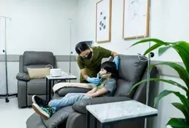 A woman in a mask and gloves gently holding a child's face indoors, sitting on a couch with furniture and decor visible in the background.