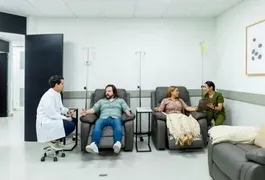 A group of people sitting in chairs in a living room, with a couch and artwork on the walls. The individuals are dressed in various clothing styles, and there is furniture scattered around the floor.