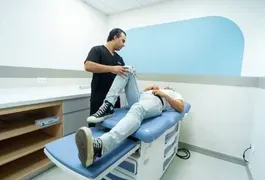 A man wearing a black shirt and black pants is lying on a blue medical bed inside a healthcare room. The setting includes various pieces of medical equipment and clean furnishings.