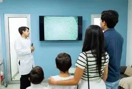 A group of people, including men, women, and a boy, are standing indoors in a room while looking at a television on the wall. They are dressed in various clothing.