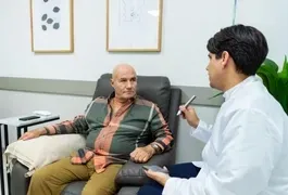 A man sitting in a chair inside a room, engaged in conversation with a doctor. The scene includes furniture and indoor elements such as walls.