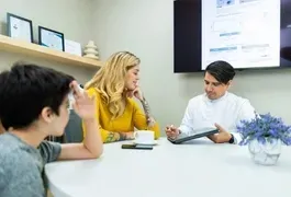 A diverse group of people sitting together at a table in an office setting, engaged in discussion or collaborative work. There is a whiteboard and various items related to education and employment around them.
