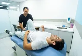 A man lying on a blue table in a medical room, with a doctor visible in the background among medical equipment.